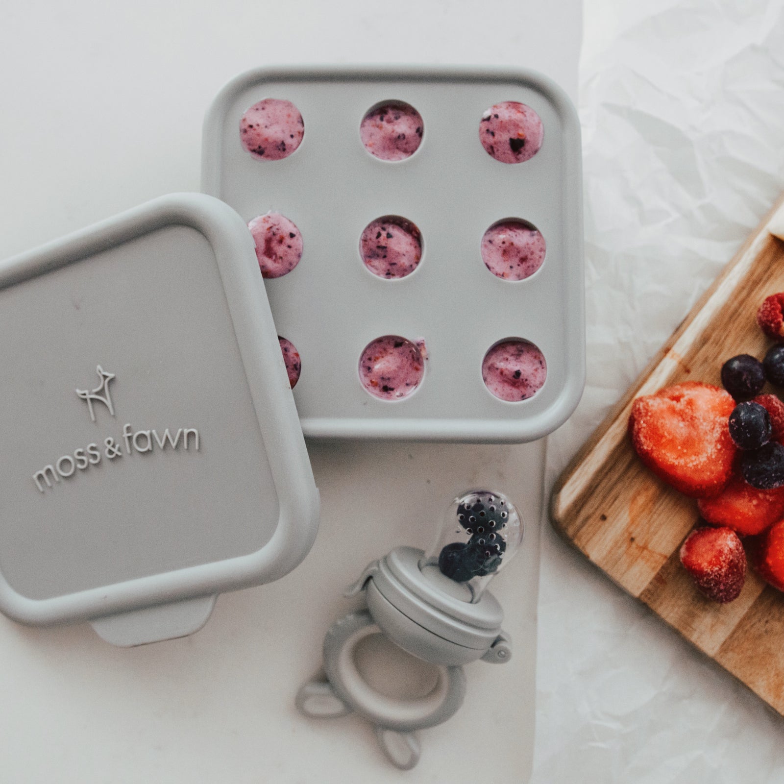 Moss & Fawn silicone food storage container with frozen berries, wooden board with fruits, and baby teether on a light surface.
