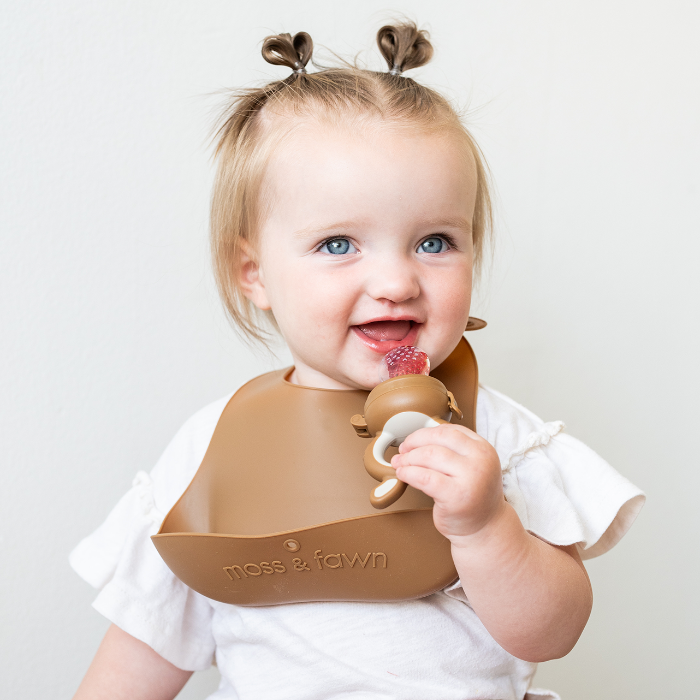 Child wearing a Moss & Fawn silicone bib and holding a forage baby food feeder filled with strawberry