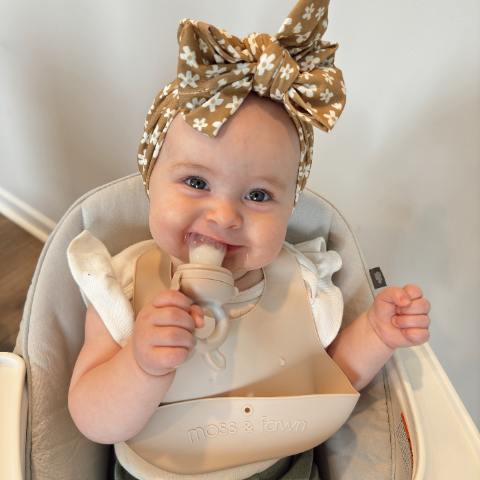 Baby in a high chair wearing a floral headband and holding a Forage Baby Food Feeder